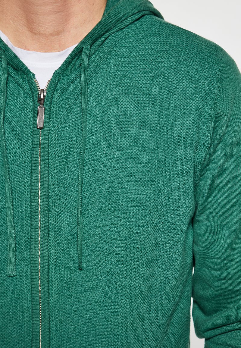 Grüner Zip-Hoodie mit strukturiertem Muster, verstellbarem Kapuzenzug und silbernem Reißverschluss.