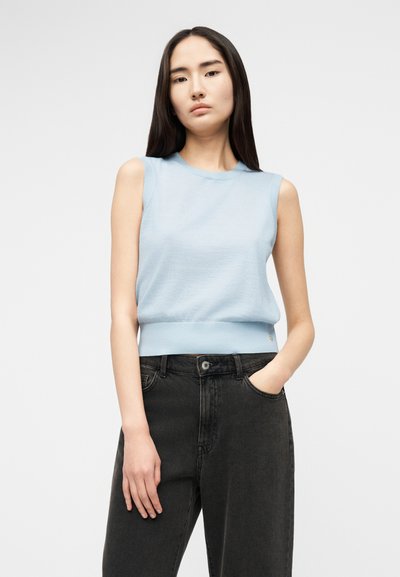 Junge Frau mit langen dunklen Haaren trägt ein ärmelloses hellblaues Oberteil und schwarze, hoch taillierte Jeans, steht mit einer Hand in der Tasche.