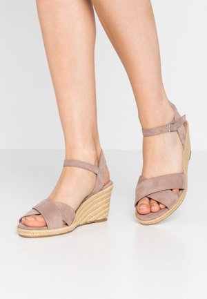 Sandały na koturnie w jasnobeżowym zamszu. Charakteryzują się krzyżującym się designem pasków oraz zapięciem w kostce. Koturn owinięty jutą, model z odkrytymi palcami.