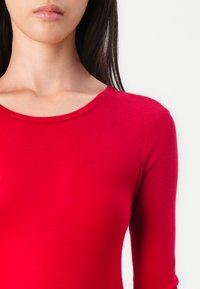 Red long-sleeve top made of ribbed fabric, featuring a round neckline and fitted design. Smooth texture with no visible accents or patterns.