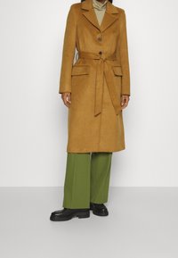 Brun yllerkappa med skärp i midjan, uppslagna slag och knappstängning. Har två framfickor. Matchas med gröna vida byxor och svarta stövlar.