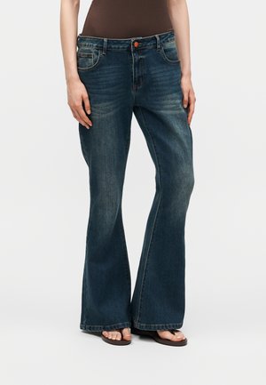 Kvinde iført mørkeblå, vide denimjeans, brun ærmeløs top og brune sandaler med åben tå, står foran en ensfarvet baggrund.