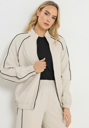 Blonde Frau trägt beigen Trainingsanzug mit schwarzer Paspelierung, schwarzes Shirt darunter, steht vor weißem Hintergrund und hält den Reißverschluss der Jacke.