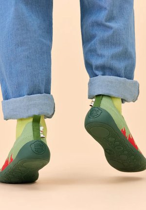 Füße in grün-roten Gummisohlen-Pantoffeln mit gelben Socken und hochgekrempelten blauen Jeans, von hinten bei einem halben Schritt gezeigt.