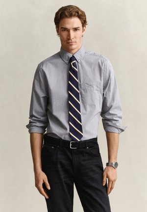 Homme portant une chemise bleu clair à boutons, une cravate rayée bleu marine, un pantalon noir et une montre argentée, debout devant un fond beige uni.