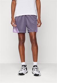Persoon staat in witte sportschoenen, witte enkelsokken, paarse sportshorts en een wit T-shirt tegen een neutrale achtergrond.