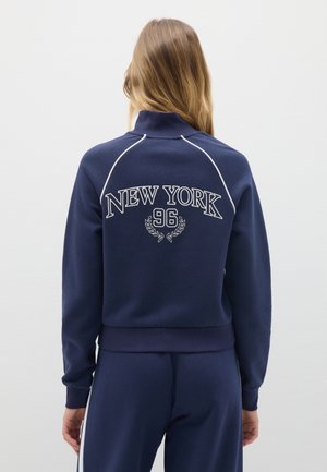 Sudadera azul marino con cuello alto, costuras blancas, "NEW YORK 96" bordado en la parte posterior, y puños y bajo acanalados.