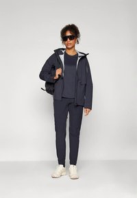 Γυναίκα με σκούρο ναυτικό μπουφάν, παντελόνι, μαύρα γυαλιά ηλίου, λευκά αθλητικά παπούτσια, κρατώντας μαύρο σακίδιο, στέκεται σε έναν απλό ανοιχτό γκρίζο τοίχο.