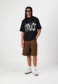 Svart grafisk t-shirt med abstrakt vitt mönster, bruna cargo-shorts, vita sneakers med grå detaljer och svarta solglasögon.