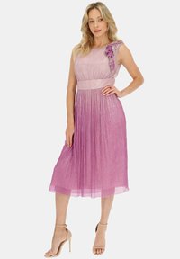 Fioletowa sukienka w gradient z jednym falbanowym ramieniem, metaliczną teksturą i plisowanym dołem, zestawiona z beżowymi sandałami na obcasie.