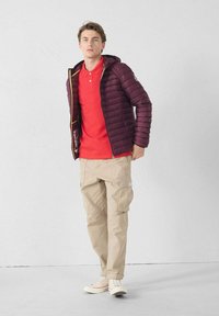 Bordeauxrode pufferjas met capuchon, rode poloshirt, kaki cargobroek en witte sneakers. De jas heeft een ritssluiting en een contrasterende voering.