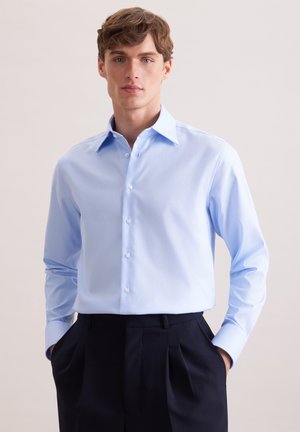 Ung mand iført en lyseblå skjorte stukket ned i sorte, plisserede bukser, stående med hænderne i lommerne mod en ensfarvet baggrund.