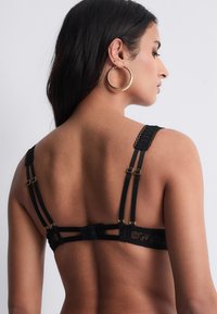 Soutien-gorge en dentelle noire avec des détails de bretelles intriqués, des bretelles réglables et des finitions en métal doré. Le dos est doté d'une fermeture à patte.