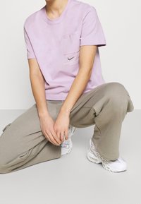 Ljuslila bomulls-T-shirt med en ficka, rund hals och Nike-logotyp. Kombinerad med lösa beige byxor och vita sportiga sneakers.