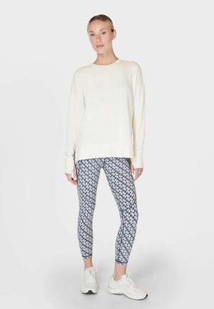 Vrouw staand, gekleed in een effen lichtcrème lange mouwen top, legging met cirkelpatroon en witte sneakers, tegen een neutrale achtergrond.