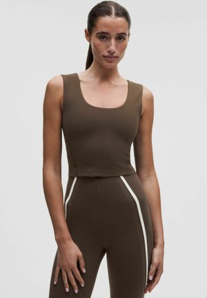 Femme aux cheveux foncés attachés en queue de cheval, portant un crop top marron sans manches et des leggings taille haute assortis avec des bandes blanches sur les côtés.