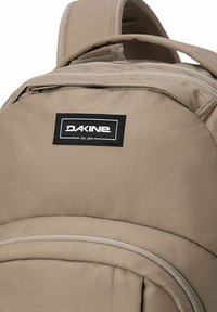 Reppure beige valmistettu kestävästä kankaasta, jossa on yläkahva, pehmustetut hihnat ja musta logo-patch. Vetoketjukiinnitys teksturoiduilla yksityiskohdilla.