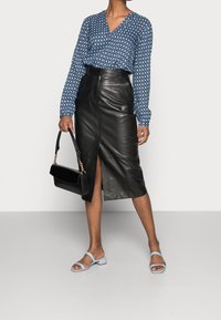 Blusa blu a fantasia con scollatura a V, gonna in pelle nera con spacco frontale e sandali blu. Tiene in mano una borsa nera con dettagli dorati.