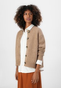Beige strikket cardigan med fem knapper, med ribbede mansjetter og en avslappet passform. Brukt over en hvit skjorte med krave og oransje skjørt.