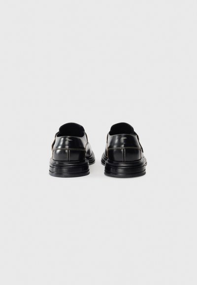 Une paire de chaussures de ville en cuir noir vues de l'arrière, montrant les détails de couture et les semelles épaisses en caoutchouc sur un fond blanc uni.