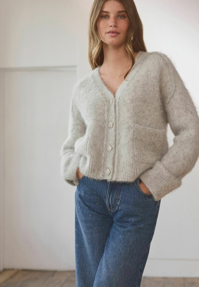 Gray, fluffy cardigan with a V-neck, five decorative buttons, and a front pocket, paired with blue jeans featuring subtle embellishments.