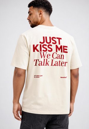 Man with short curly hair turns back, wearing beige T-shirt with red text "JUST KISS ME We Can Talk Later" and smaller inscriptions.