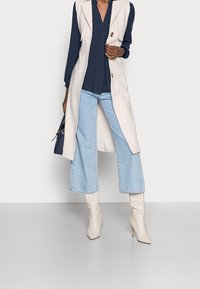 Woman wearing a beige sleeveless coat over a navy blouse, light blue wide-leg jeans, white heeled boots, and holding a navy handbag.