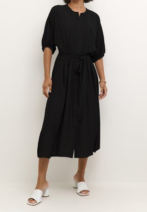 Femme portant une robe midi noire boutonnée avec une taille nouée, des manches courtes bouffantes et des sandales blanches à talons ouvertes.