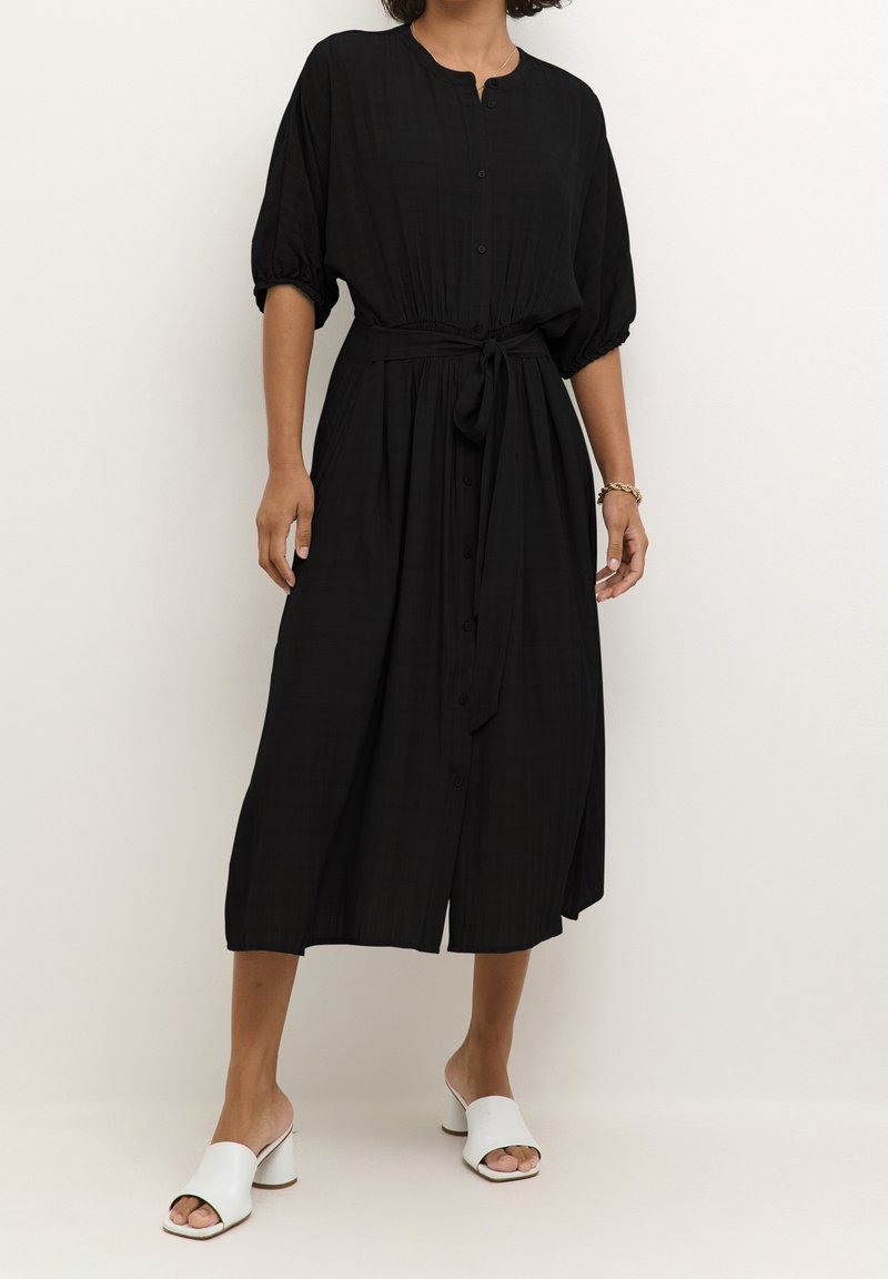 Femme portant une robe midi noire boutonnée avec une taille nouée, des manches courtes bouffantes et des sandales blanches à talons ouvertes.