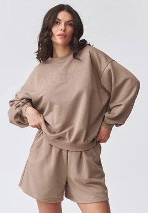 Kobieta o ciemnych falowanych włosach ma na sobie luźną beżową bluzę i dopasowane szorty, pozując z rękami opartymi na biodrach na jednolitym tle.