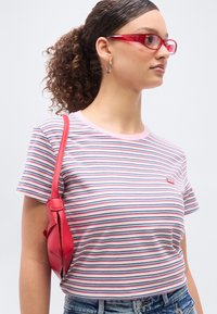 Νεαρή γυναίκα με σγουρά μαλλιά σε αλογοουρά, φοράει κόκκινα γυαλιά και ριγέ μπλουζάκι, κρατώντας μια  μικρή κόκκινη τσάντα ώμου, κοιτάζοντας προς τα δεξιά.