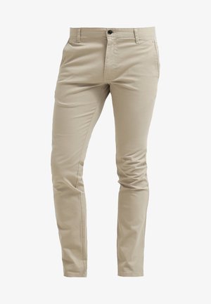 Marineblauer Trenchcoat mit Schulterklappen, weißes Hemd, beige Chinos und schwarze Stiefeletten mit strukturierten Sohlen, in einer dynamischen Pose stehend.