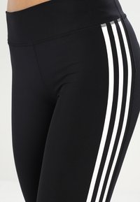 Leggings negros hechos de un material elástico que presentan tres rayas blancas verticales a lo largo del costado exterior y una cinturilla alta.