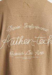 Ruda medžiaga su balta siuvinėta užrašu: "Chasin' Performance Authen-tech Research Lab Ams" su geometrišku logotipu viršuje.