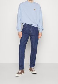 Světle modrá mikina s malým zlatým logem, skloubená s tmavě modrými džínami a hnědými teniskami s bílou podrážkou. Neformální outfit.