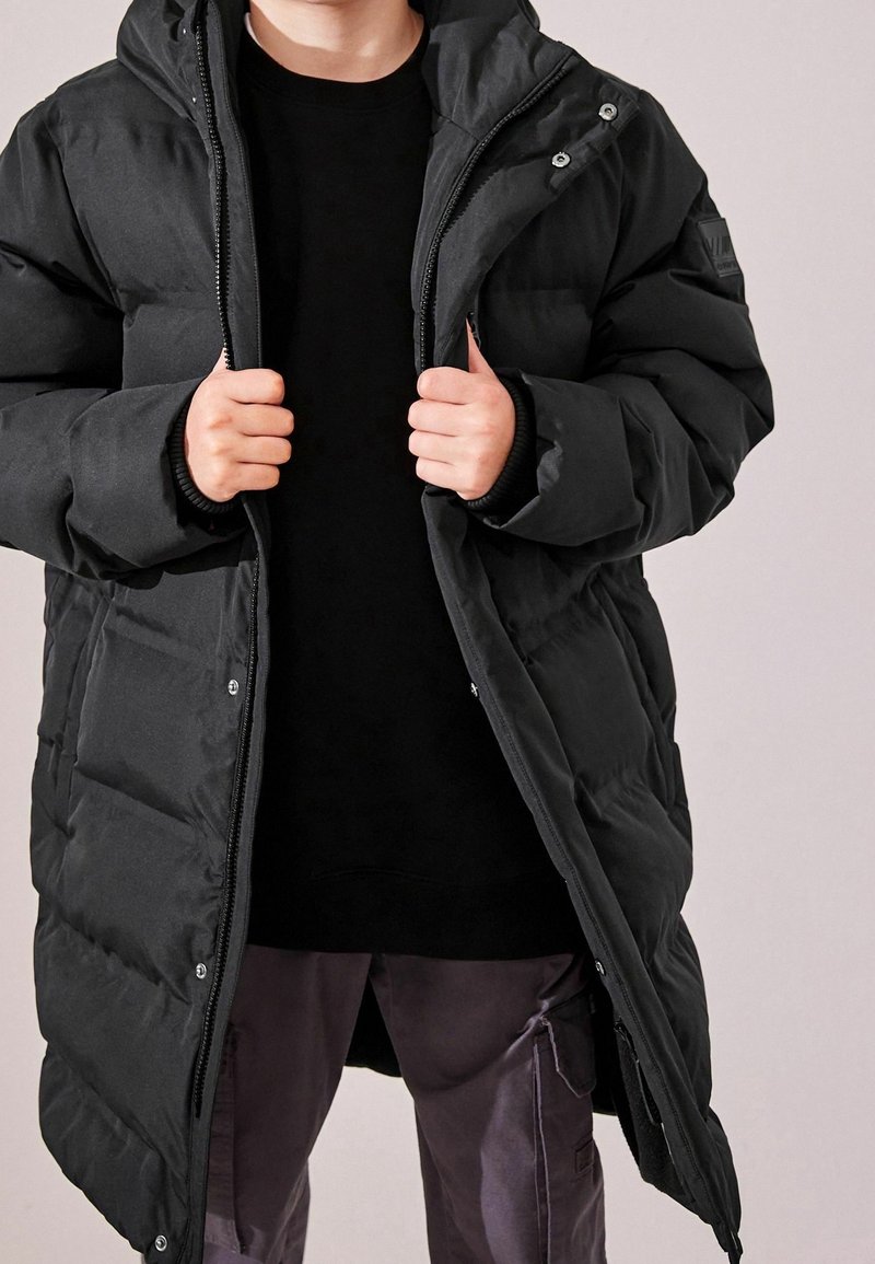 Enfant portant une longue doudoune noire avec capuche, partiellement dézippée, et un pantalon cargo foncé, sur un fond uni.