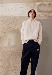 Light beige long-sleeve button-up shirt and dark navy wide-leg pants with a red and black Nike logo on the left thigh. Smooth texture.