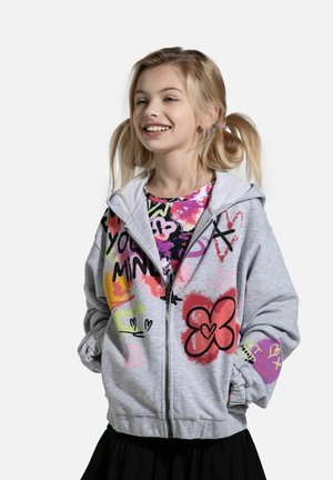 Szara bluza z zamkiem, zdobiona kolorowymi wzorami w stylu graffiti i o miękkiej fakturze. Zakrywa wzorzysty T-shirt, znajdujący się pod nią, i jest zestawiona z czarną spódnicą.