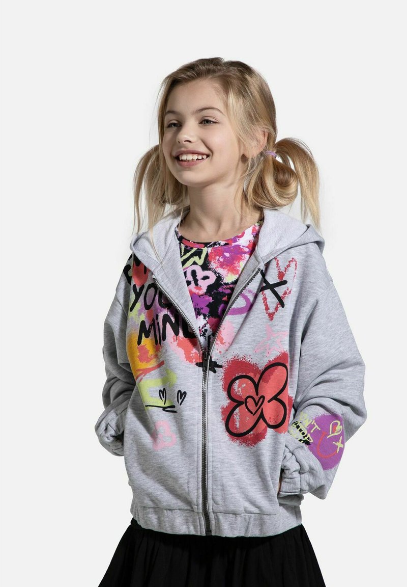 Szara bluza z zamkiem, zdobiona kolorowymi wzorami w stylu graffiti i o miękkiej fakturze. Zakrywa wzorzysty T-shirt, znajdujący się pod nią, i jest zestawiona z czarną spódnicą.