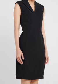 Robe noire sans manches avec un col haut et plié, et une silhouette ajustée. Tissu lisse avec des détails de couture en diagonale et une longueur jusqu'aux genoux.