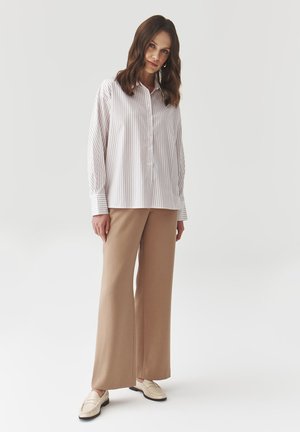 Gestreifte, knöpfbare Bluse in Weiß und Hellrose, kombiniert mit eleganten, weiten beigen Hosen und beigen Slippern vor einem schlichten Hintergrund.