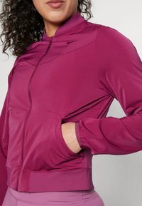 Mujer con una chaqueta de cremallera magenta, con la mano en el bolsillo, que muestra puños y cuello acanalados, y un pequeño logo de Nike en el pecho.