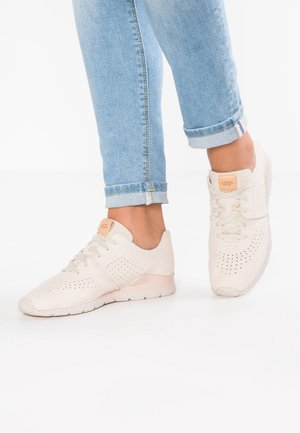 Stopy w jasnoniebieskich, podwiniętych dżinsach noszące kremowe buty sportowe UGG z perforowanymi detalami i beżowymi logo na białym tle.