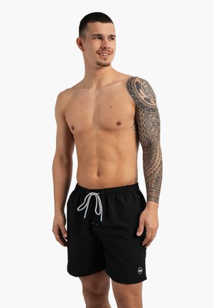 Jeune homme avec un tatouage sur le bras, portant un short de bain noir avec un cordon blanc, debout et souriant légèrement en regardant vers la droite.