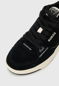Black suede sneaker with a white rubber sole. Features textured stitching, a triangular accent, and the "GUESS" logo on the side.