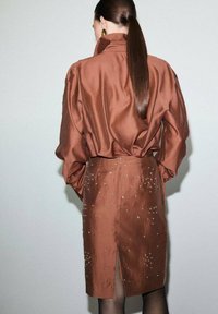 Vestido camisero de satén marrón con cuello alto, de corte holgado y abertura frontal. Decorado con tachuelas metálicas. Cabello largo y liso a la vista.
