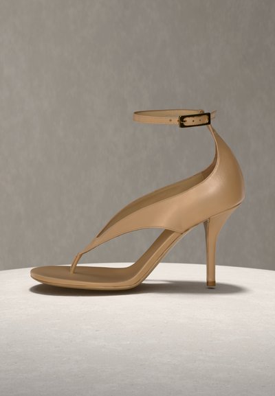 Μπεζ ψηλοτάκουνο σανδάλι με λουράκι στον αστράγαλο και σχεδιασμό με μυτερό λουράκι ανάμεσα στα δάχτυλα, τοποθετημένο πάνω σε λεία, ανοιχτή επιφάνεια με ουδέτερο γκρι φόντο.