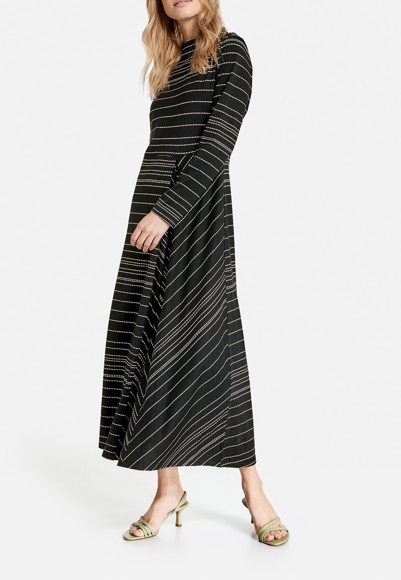 Femme portant une robe maxi noire avec des rayures horizontales et diagonales beiges, associée à des sandales à talons à lanières vert clair.