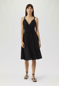 Schwarzes Kleid mit einem gehäkelten Oberteil und tiefem V-Ausschnitt; fließender Rock, der bis zur Wade reicht; kombiniert mit schwarzen Riemchensandalen.
