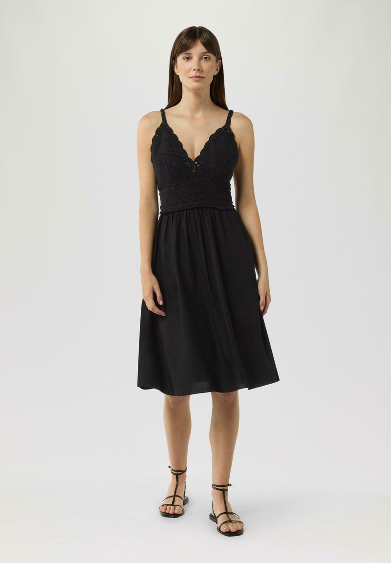 Schwarzes Kleid mit einem gehäkelten Oberteil und tiefem V-Ausschnitt; fließender Rock, der bis zur Wade reicht; kombiniert mit schwarzen Riemchensandalen.